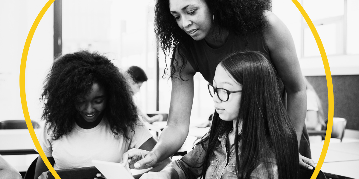 A Black female teacher assists two middle school girls, one Black and one Asian, in a classroom setting. The image is black and white with a yellow circle partially framing the scene.