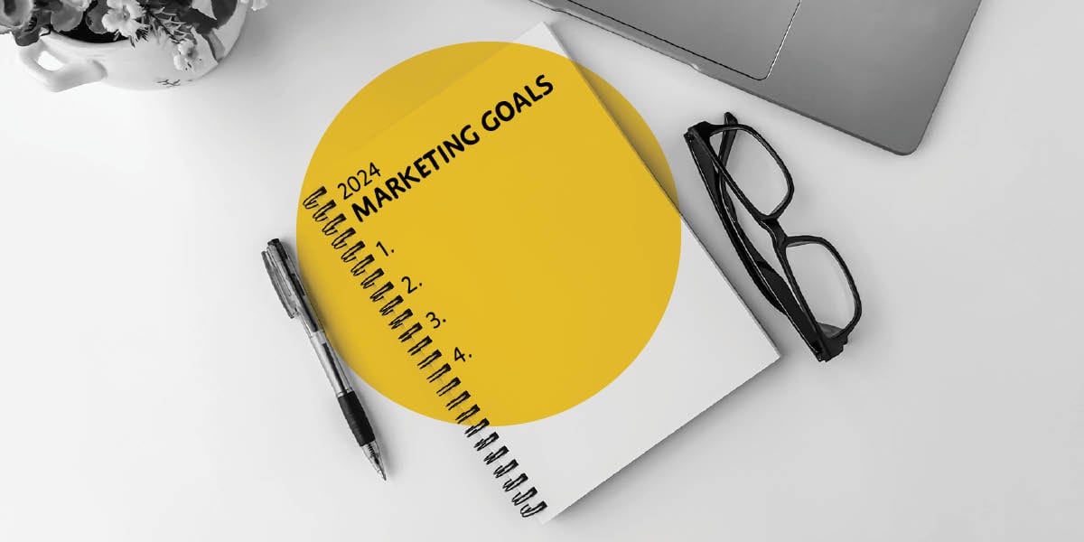 A spiral-bound notebook with 4 marketing goals on it, flanked by a pen and glasses